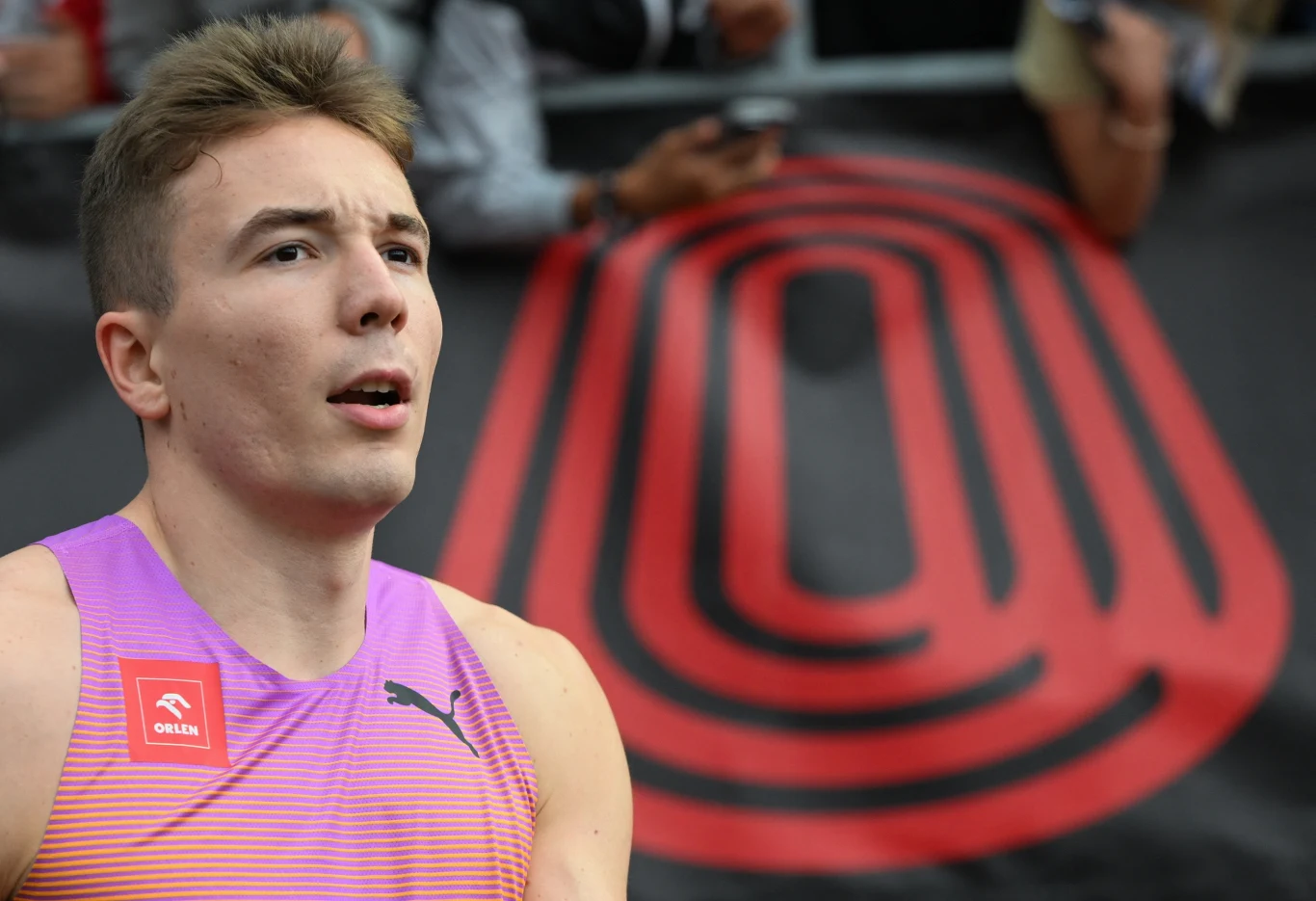 Młody sportowiec w stroju do biegania na tle dużego logotypu lub symbolu na stadionie, w otoczeniu kibiców.