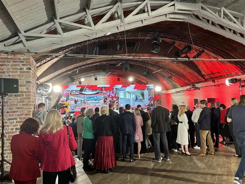 Wyniki late poll. Radość w sztabie PiS Grupa ludzi zgromadzona w hali z cegieł i stalowych konstrukcji, obserwująca prezentację na scenie z dużymi ekranami, oświetlenie sceniczne, czerwona iluminacja na ścianach.