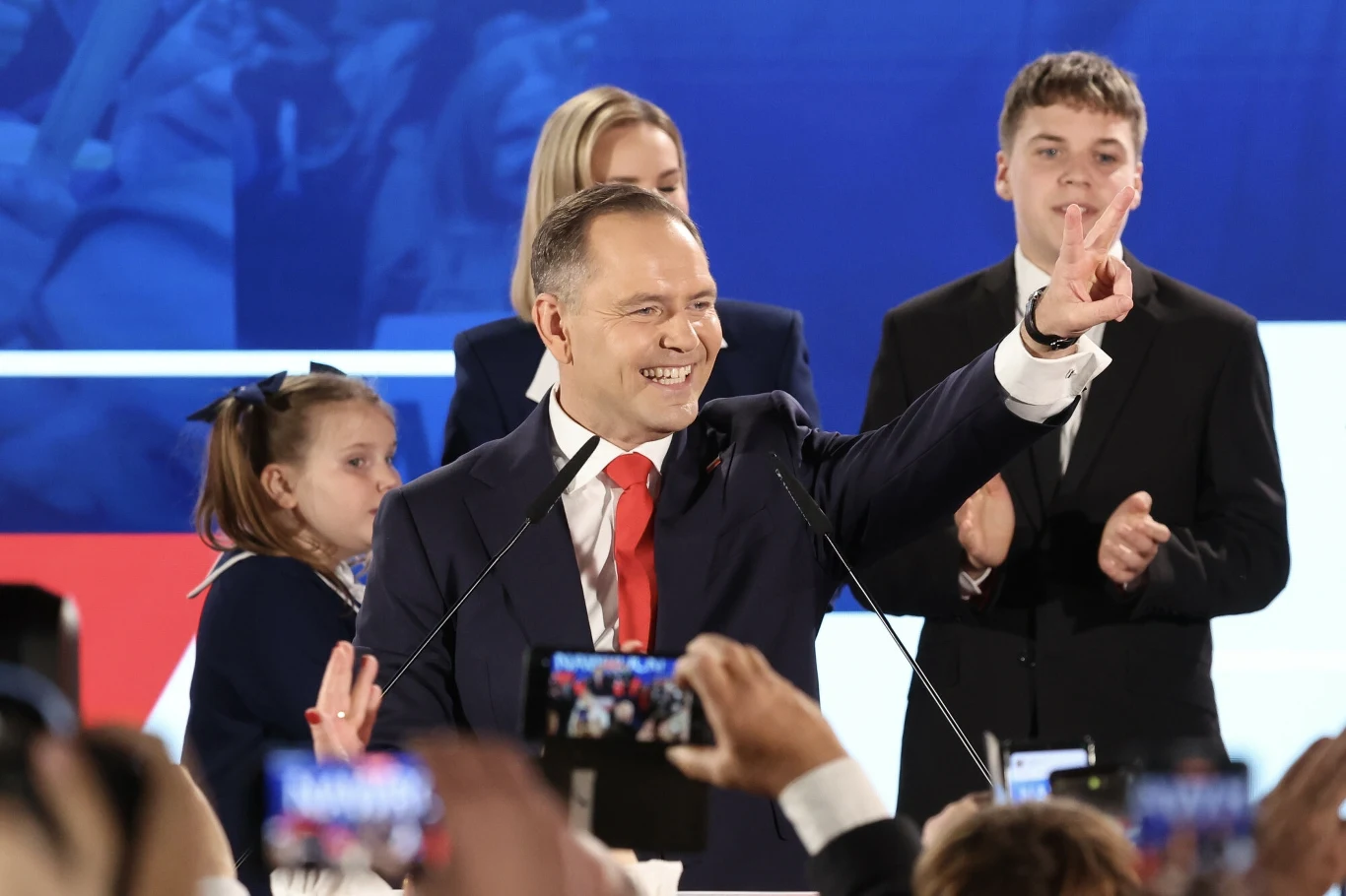 Karol Nawrocki po ogłoszeniu wyników exit poll Mężczyzna ubrany w garnitur i czerwoną krawat przemawia do tłumu, wykonując gest zwycięstwa dłońmi, za nim stoją cztery osoby, w tym dzieci, a przed sceną znajdują się osoby fotografujące go smartfonami.