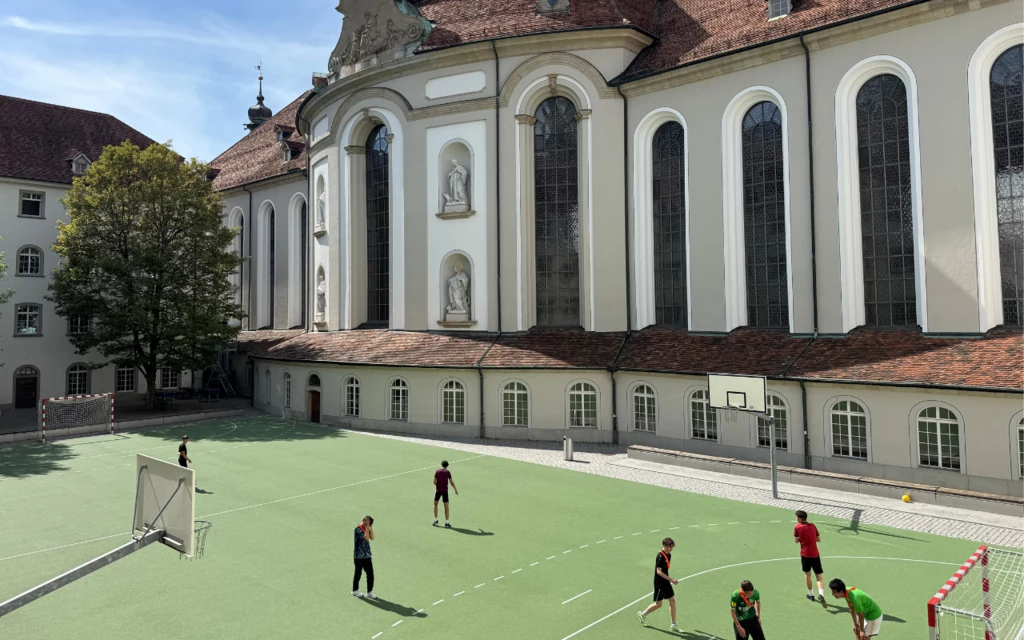 Pełnowymiarowe boisko na dziedzińcu opactwa w Sankt Gallen Grupa dzieci i młodzieży gra w piłkę nożną na zielonym boisku przy dużym, zabytkowym budynku z wysokimi oknami i barokowymi zdobieniami, w tle widoczne fragmenty drzew i architektura kościoła.