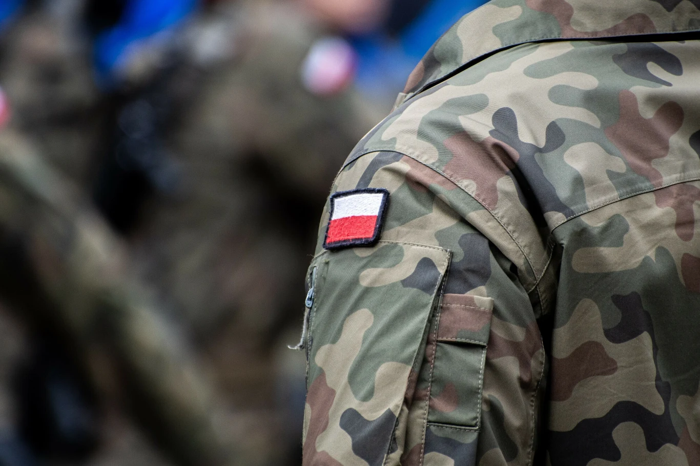 Żołnierz w większości poparli w wyborach Karola Nawrockiego (zdjęcie ilustracyjne) Mundur wojskowy z naszywką flagi Polski na ramieniu, motyw wojskowego kamuflażu, w tle rozmyte sylwetki innych osób w podobnych mundurach.