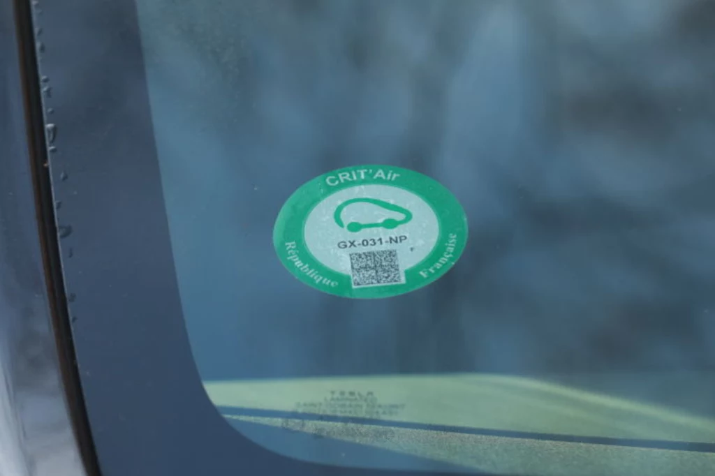 Winieta Crit'Air zezwalała na wjazd do obszarów objętych zakazem. Jej brak wiąże się z mandatem w wysokości do 135 euro. Naklejka ekologiczna Crit'Air umieszczona w lewym dolnym rogu szyby samochodu, jasna zielona etykieta z symbolem auta i kodem QR, tło lekko rozmyte.