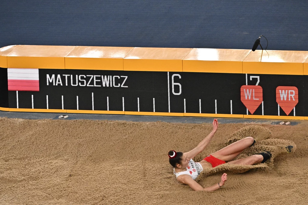 Anna Matuszewicz Atletka ubrana w biało-czerwony strój lądująca w piaskownicy podczas skoku w dal, w tle tablica z nazwiskiem zawodniczki i oznaczeniem odległości oraz flagą Polski.