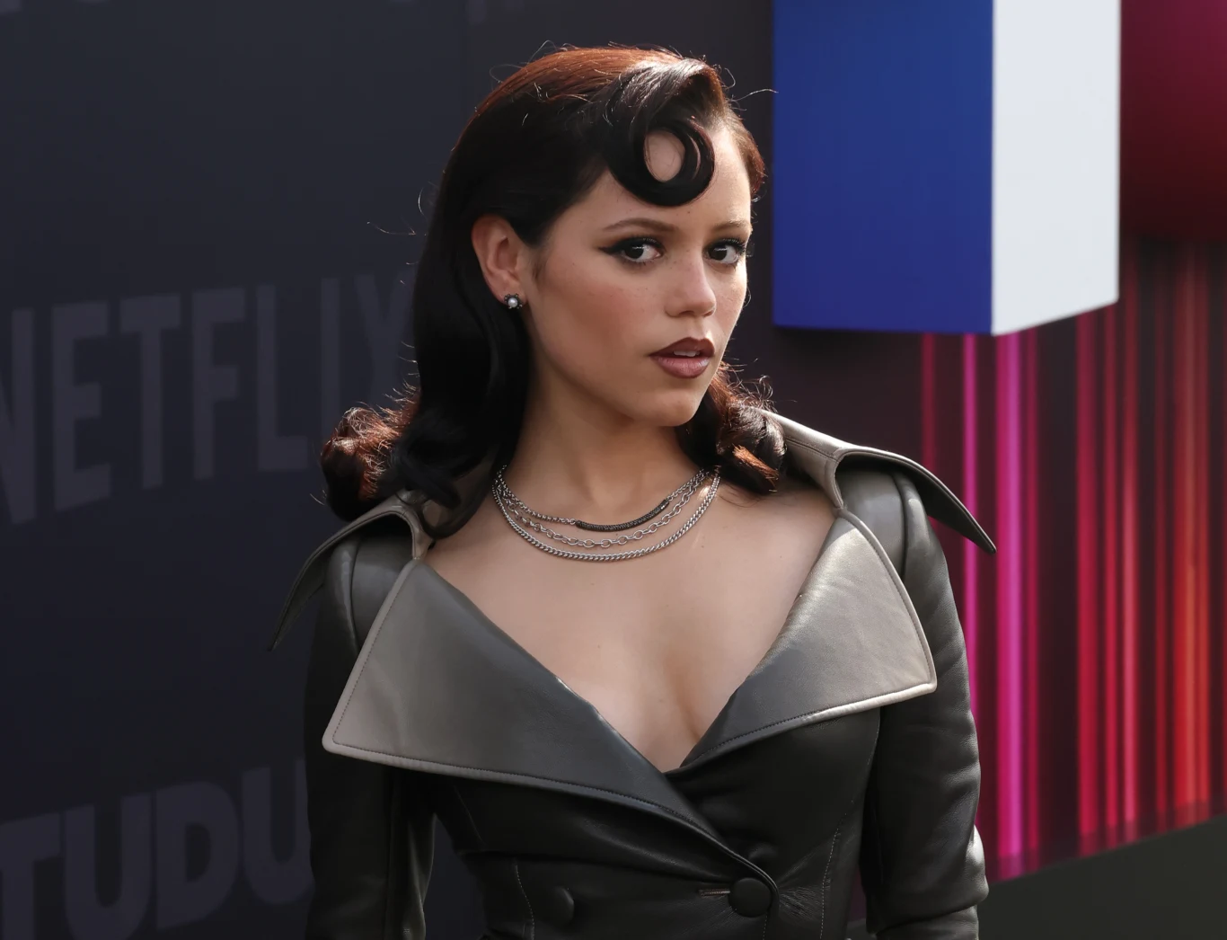 Jenna Ortega zachwyciła kreacją Kobieta o ciemnych, falowanych włosach, ubrana w ciemny płaszcz ze skórzanym kołnierzem i głębokim dekoltem, ma na szyi elegancki naszyjnik, stoi na tle napisu NETFLIX oraz kolorowej dekoracji geometrycznej.