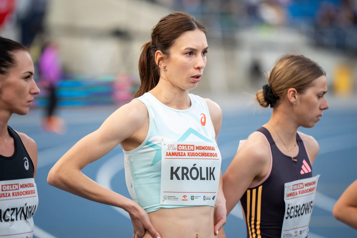 Trzy zawodniczki przygotowujące się do startu w biegu na stadionie lekkoatletycznym, każda z numerem startowym i nazwiskiem na koszulce, w tle rozmyte trybuny.