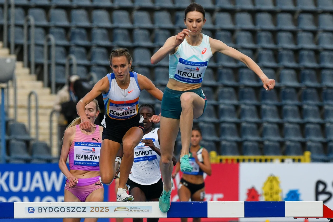 Zawodniczki biegnące w biegu z przeszkodami podczas zawodów lekkoatletycznych, przeskakujące przez płotek na stadionie z pustymi trybunami w tle.