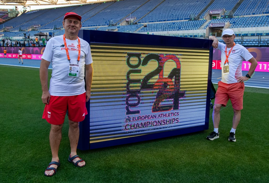 Jacek Kostrzeba (po prawej) razem ze Stanisławem Jaszczakiem Dwóch mężczyzn w sportowych strojach i czapkach pozuje przy dużym ekranie LED wyświetlającym logo Mistrzostw Europy w Lekkoatletyce 2024 na tle pustych trybun stadionu.
