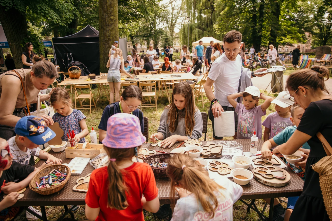 Grupa dzieci i dorosłych siedzi przy stole na świeżym powietrzu, uczestnicząc w warsztatach plastycznych, podczas których tworzą prace rękodzielnicze z drewna, wokół panuje atmosfera pikniku rodzinnego.
