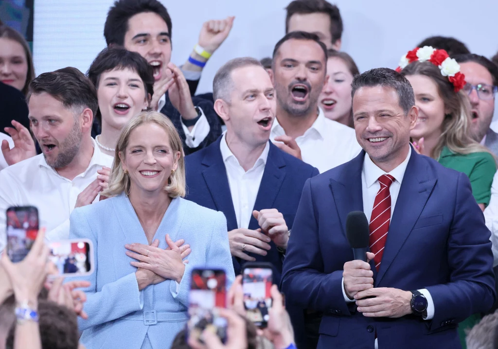 Wieczór wyborczy Rafała Trzaskowskiego Grupa osób w eleganckich ubraniach, uśmiechnięci, zgromadzeni na scenie i celebrujący wydarzenie publiczne, na pierwszym planie kobieta trzymająca dłonie na sercu i mężczyzna w garniturze z mikrofonem, w tle tłum ludzi robiących zdjęcia telefonami.