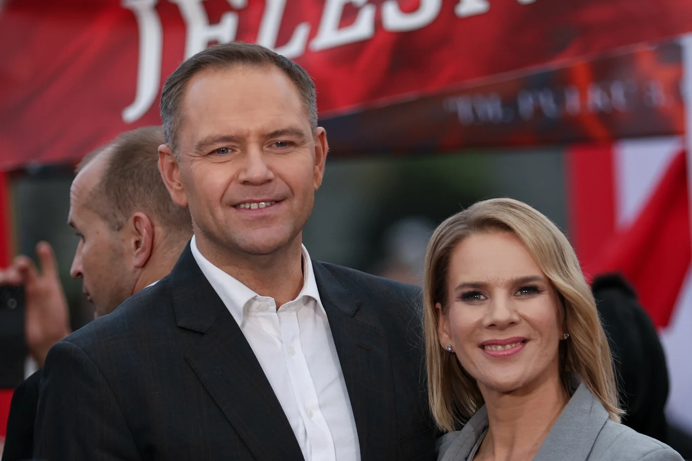 Uśmiechnięta para w eleganckich ubraniach pozuje razem na tle czerwono-białego baneru.