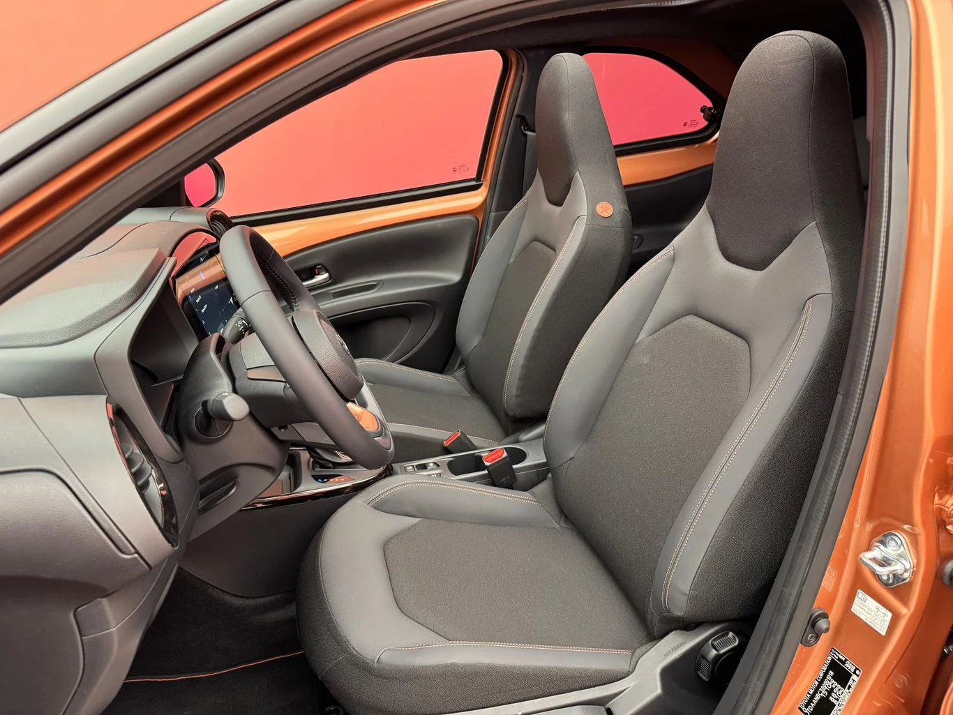 Wewnątrz nowoczesnego samochodu znajdują się dwa komfortowe fotele przednie, deska rozdzielcza z panelem sterowania oraz elegancka kierownica, całość utrzymana w czarno-pomarańczowej kolorystyce.