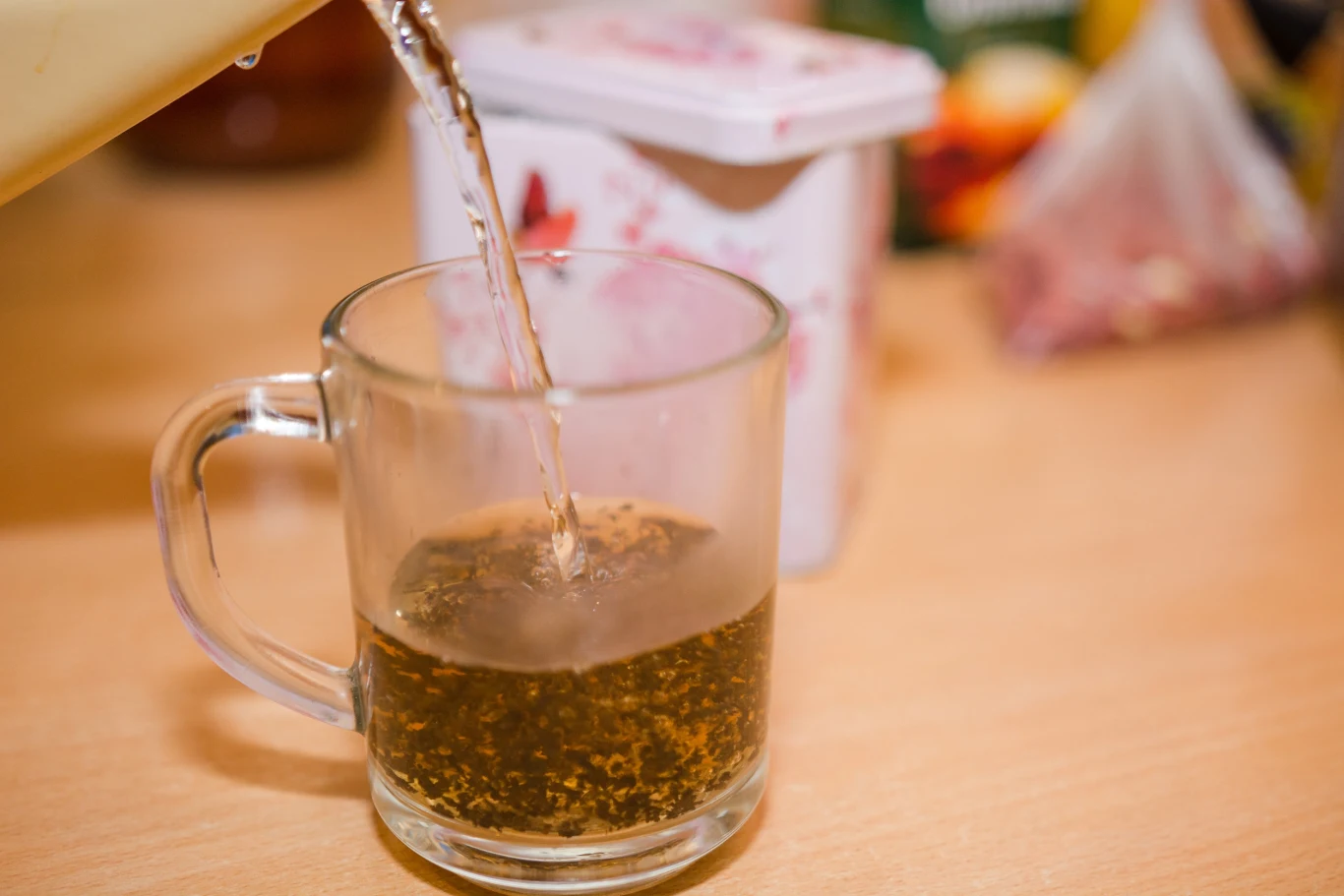 Szklana filiżanka z przezroczystym uchem, do której wlewana jest gorąca woda, a liście herbaty znajdują się na dnie, w tle rozmazane opakowanie i produkty spożywcze na blacie kuchennym.