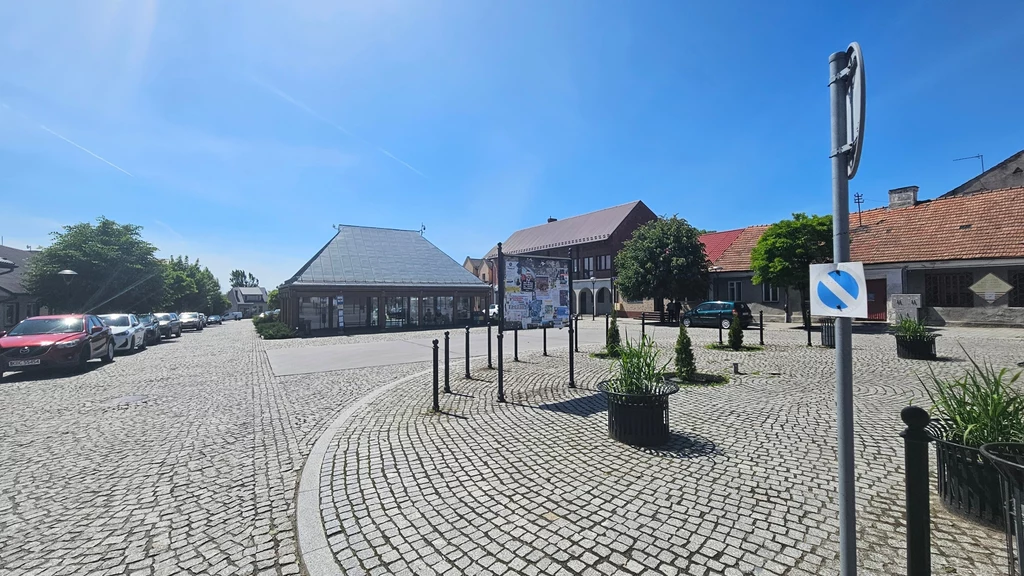 Rynek w Nowym Brzesku Puste brukowane skrzyżowanie w centrum małego miasta, kilka zabytkowych budynków z czerwonymi i grafitowymi dachami, po lewej stronie zaparkowane samochody, w centrum placu zielone donice, znak drogowy zakazujący zatrzymywania się, jasne niebo bez chmur.