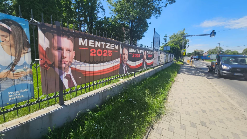 Plakaty wyborcze polityka z napisem Mentzen 2025 rozwieszone na ogrodzeniu wzdłuż chodnika, po lewej stronie widoczny fragment innej reklamy z kobietą, po prawej ulicą przejeżdża samochód, w tle drzewa i niebieskie niebo.