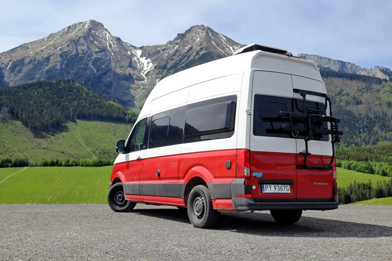 Volkswagen Grand California to największy kamper w gamie producenta. Polska to dla niego bardzo ważny rynek. Czerwono-biały kamper zaparkowany na tle gór z ośnieżonymi szczytami, wokół zielone pola i lasy, atmosfera wypoczynku i podróży.