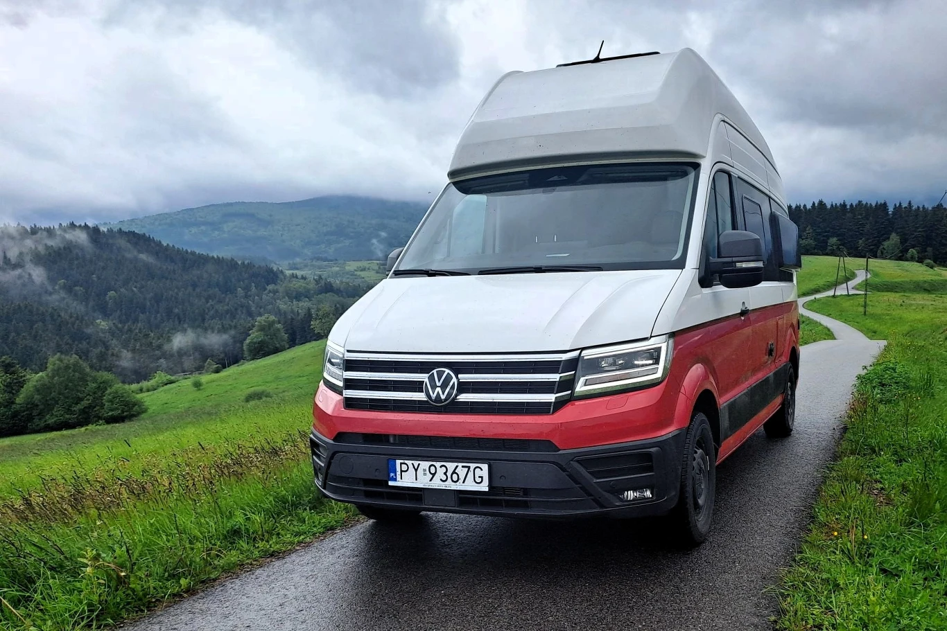 Volkswagen Grand California zbudowana jest na dostawczym Crafterze. Czerwono-biały kamper marki Volkswagen stoi na wąskiej asfaltowej drodze otoczonej zielonymi łąkami i lasami, w tle widoczne są góry częściowo przykryte chmurami.