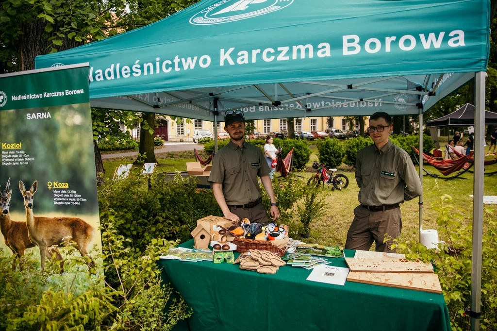 Stoisko edukacyjne Nadleśnictwa Karczma Borowa, na którym dwóch leśników prezentuje materiały edukacyjne oraz modele zwierząt i budki lęgowe, obok widoczny baner z informacjami o sarnach.