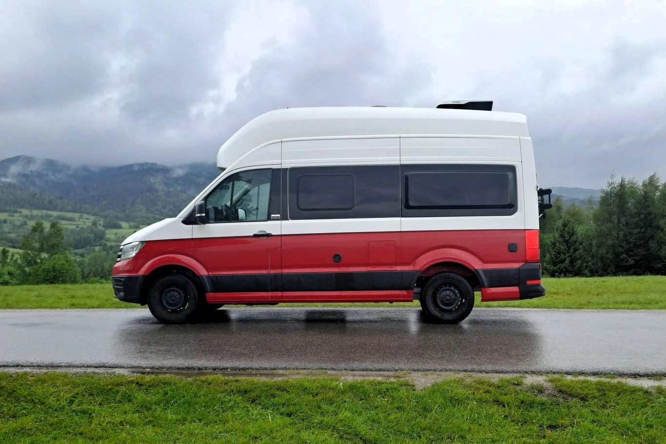 Co ciekawe Polska jest drugim rynkiem pod względem sprzedaży Volkswagena Grand Californii. Furgonetka typu kamper w biało-czerwonych barwach zaparkowana na drodze, w tle widać zielone pola, drzewa oraz górzysty krajobraz pod pochmurnym niebem.