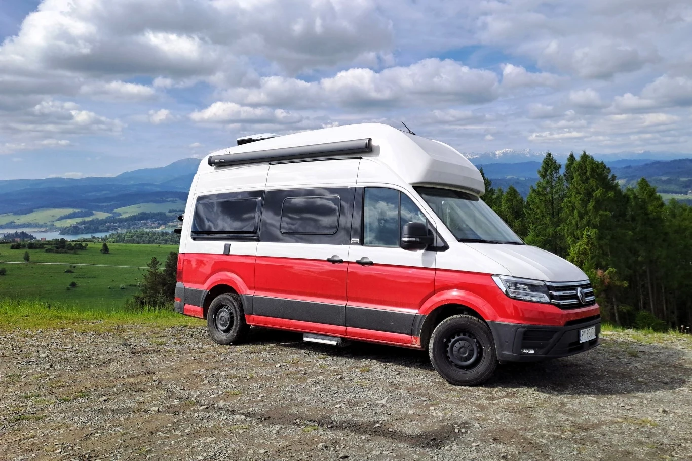 Za wprawianie w ruch Volkswagena Grand Californii odpowiada dwulitrowy silnik Diesla o mocy 163 KM. Czerwono-biały kamper zaparkowany na tle zielonych pól i gór, w słoneczny dzień, z lasem po prawej stronie i rozproszonymi chmurami na niebie.