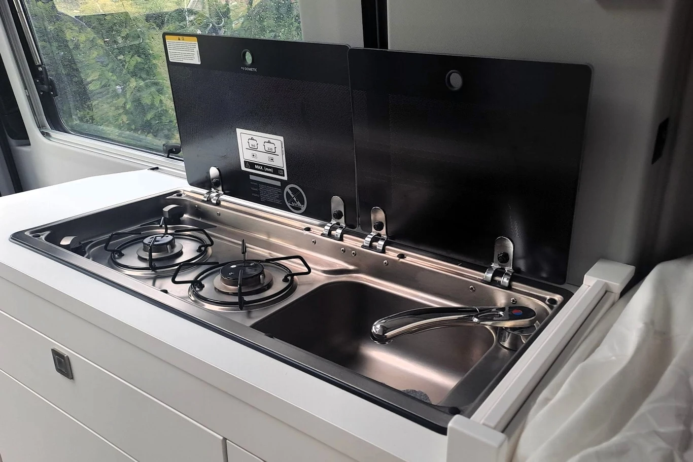 Wewnątrz Volkswagena Grand Californii znajdziemy m.in. dwupalnikową kuchenkę gazową oraz zlew. Dwupalnikowa kuchenka gazowa i stalowy zlew z baterią, zamontowane w kompaktowej zabudowie kuchennej pojazdu kempingowego, okno z widokiem na zieleń w tle
