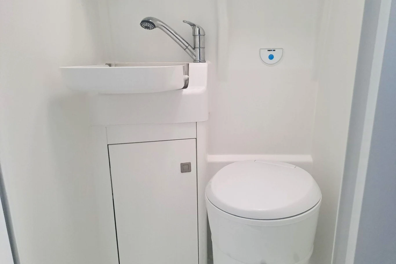 Volkswagen Grand California wyposażony jest także w toaletę z prysznicem. Niewielka, kompaktowa łazienka z białą toaletą i umywalką umieszczoną nad szafką, minimalna ilość detali, całość utrzymana w białej kolorystyce, praktyczne rozwiązanie do małych przestrzeni.