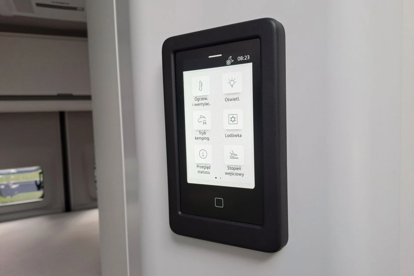 Do obsługi najważniejszych funkcji w Grand Californii służy oddzielny wyświetlacz znajdujący się w przestrzeni mieszkalnej. Można nimi sterować również z ekranu multimediów. Panel sterowania w nowoczesnym kamperze zamontowany na jasnej ścianie, wyświetlający różne opcje obsługi urządzeń wewnętrznych takich jak oświetlenie, lodówka, system ogrzewania czy tryb kempingu.