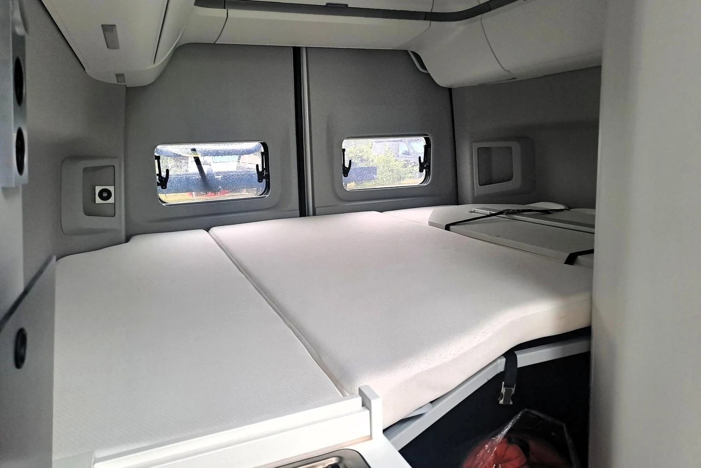 W krótszej Grand Californii mamy dwa łóżka - jedno na końcu auta, a drugie nad przestrzenią kierowcy. Wnętrze kabiny ciężarówki z rozłożoną przestrzenią do spania, widoczne dwa materace, trzy niewielkie okna po bokach i z tyłu, jasno oświetlone, z elementami schowków nad łóżkiem.