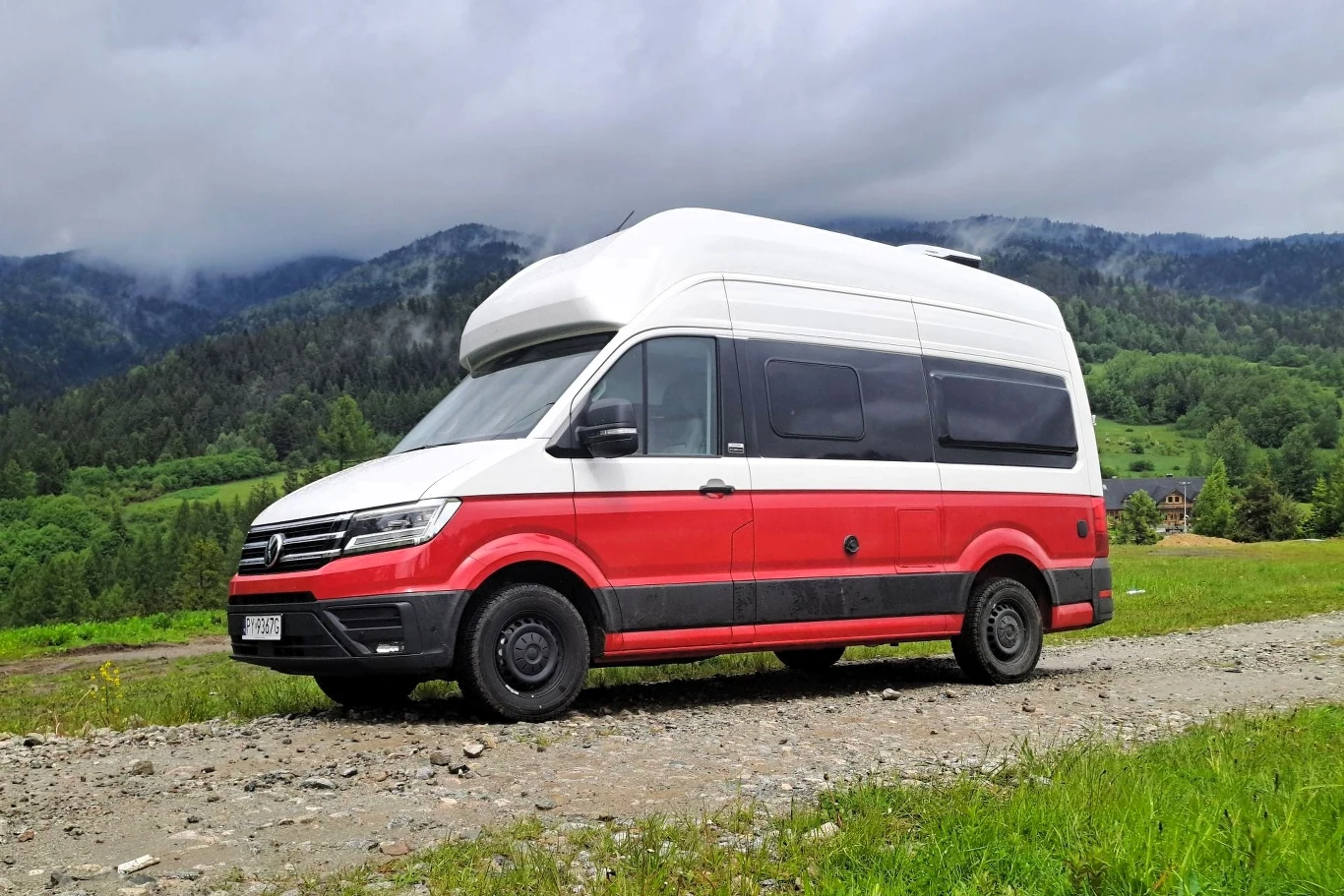 Ceny Volkswagena Grand Californii startują od 391 140 zł. Czerwono-biały kamper zaparkowany na żwirowej drodze, w tle zalesione wzgórza i górski krajobraz, częściowo pochmurne niebo