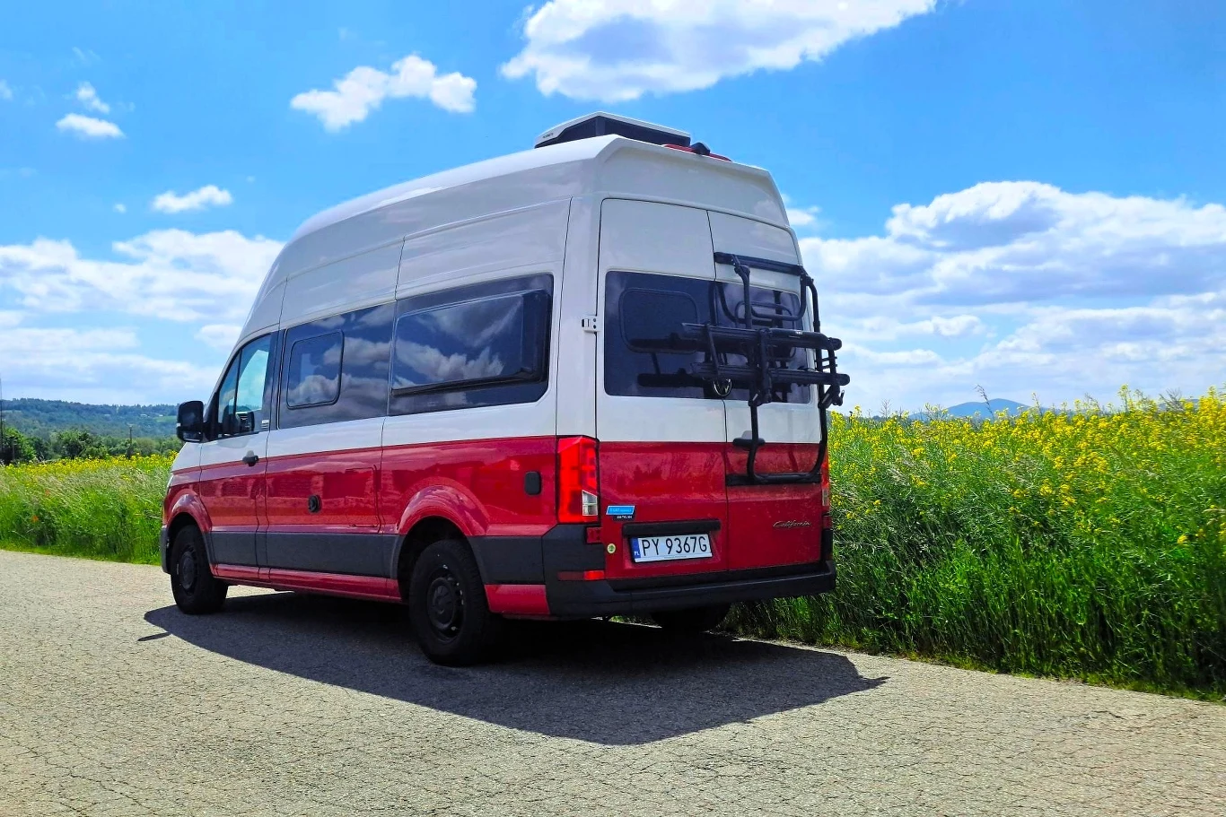 Volkswagen Grand California generuje tylko 163 KM. Moim zdaniem mogłoby być ich więcej. Czerwono-biały kamper zaparkowany przy asfaltowej drodze, otoczony zielonymi polami i żółtymi kwiatami, w tle błękitne niebo z białymi chmurami.