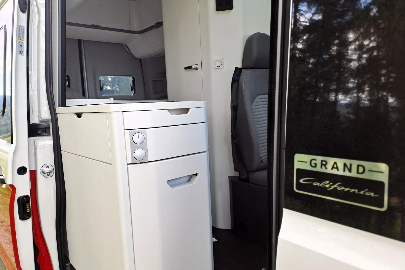 Na wyposażeniu Volkswagena Grand Californii znaleźć można m.in. lodówkę z zamrażarką. Wnętrze kampera Grand California z widocznymi białymi meblami kuchennymi oraz fragmentem siedzenia kierowcy, przez szeroko otwarte drzwi pojazdu.