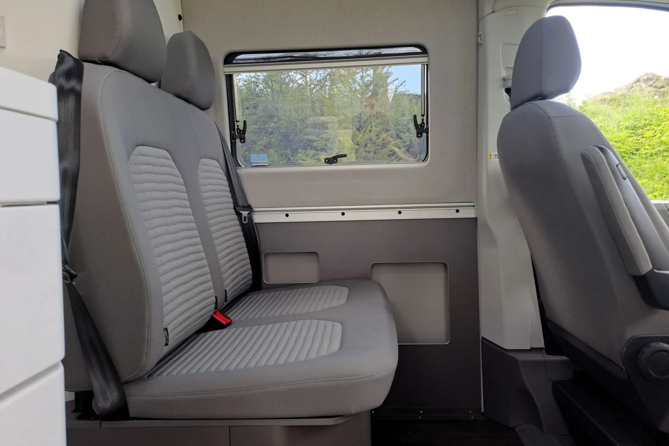 Niezależnie od wybranej wersji długości Grand California oferuje cztery miejsca. Dwie szare siedzenia w przedziale pasażerskim pojazdu, wysokie oparcia, w tle okno z widokiem na zieleń, po prawej stronie częściowo widoczne siedzisko kierowcy.