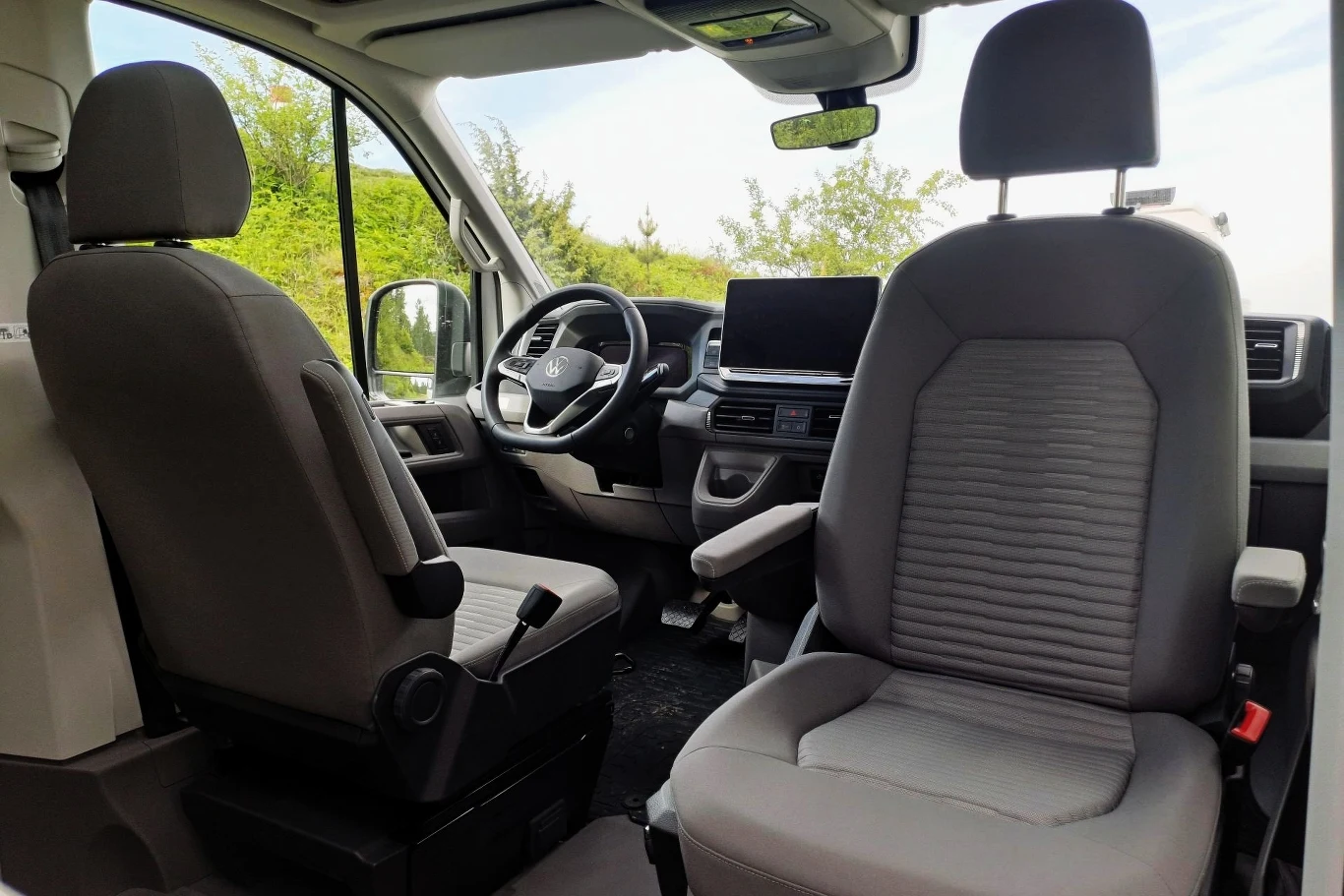 Przednie fotele w Grand Californii są obracane. Przestronne wnętrze samochodu dostawczego z dwoma siedzeniami kierowcy i pasażera, nowoczesną deską rozdzielczą, dużym wyświetlaczem centralnym oraz widokiem na zieleń przez boczne okno.