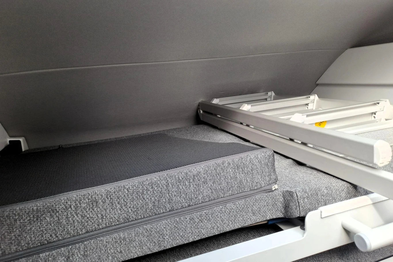 Jedno łóżko w Grand Californii znajduje się na końcu auta, drugie, rozkładane, nad przestrzenią kierowcy. Szara tapicerowana powierzchnia złożonego materaca lub łóżka w wąskim wnętrzu, obok metalowa rama mechanizmu rozkładania.