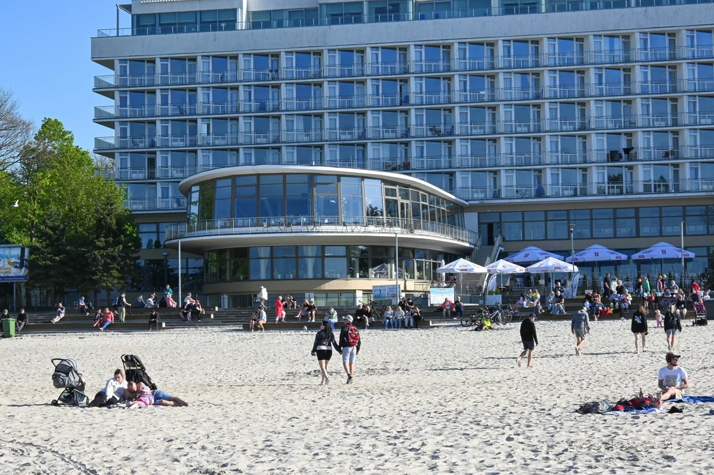 Ludzie spacerują i odpoczywają na piaszczystej plaży przed nowoczesnym, wielopiętrowym hotelem z dużymi oknami i tarasem. W tle widoczne są drzewa oraz strefa z parasolami, przy której zgromadzeni są turyści.