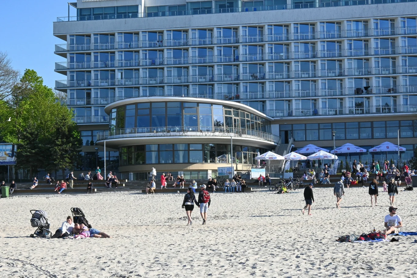 Ludzie spacerują i odpoczywają na piaszczystej plaży przed nowoczesnym, wielopiętrowym hotelem z dużymi oknami i tarasem. W tle widoczne są drzewa oraz strefa z parasolami, przy której zgromadzeni są turyści.