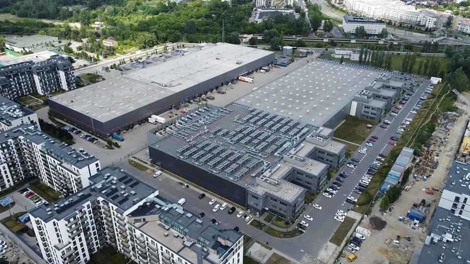 Największe centrum danych w kraju? Będzie w warszawskim Ursusie Nowoczesny kompleks magazynowo-biurowy otoczony osiedlami mieszkaniowymi, widoczna infrastruktura techniczna na dachach budynków, samochody zaparkowane wokół obiektów, zielone tereny w tle.