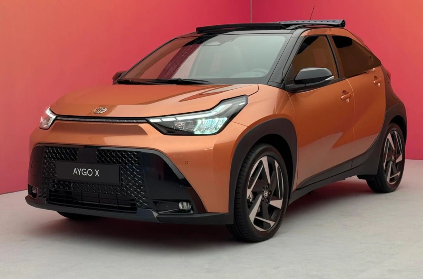 Nowoczesny, kompaktowy samochód miejski Toyota Aygo X w kolorze miedzianym, ustawiony na szarym tle z różowymi i pomarańczowymi odcieniami, z wyrazistymi reflektorami i dużymi felgami aluminiowymi.