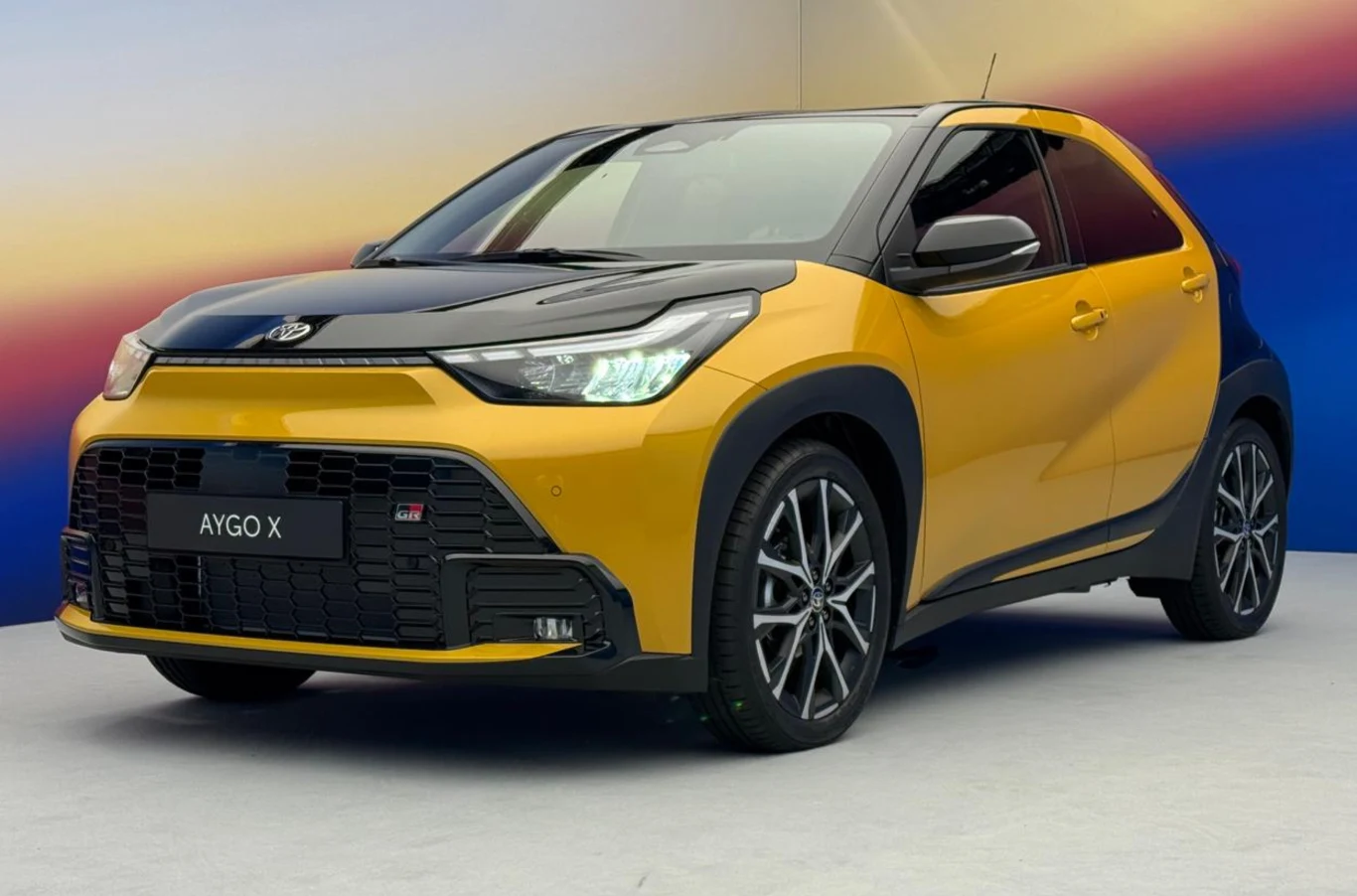 Nowoczesny, miejski samochód Toyota Aygo X w dwukolorowym żółto-czarnym nadwoziu, sportowe felgi, charakterystyczny grill oraz designerskie reflektory LED, auto ustawione na jasnym tle z gradientem kolorystycznym.