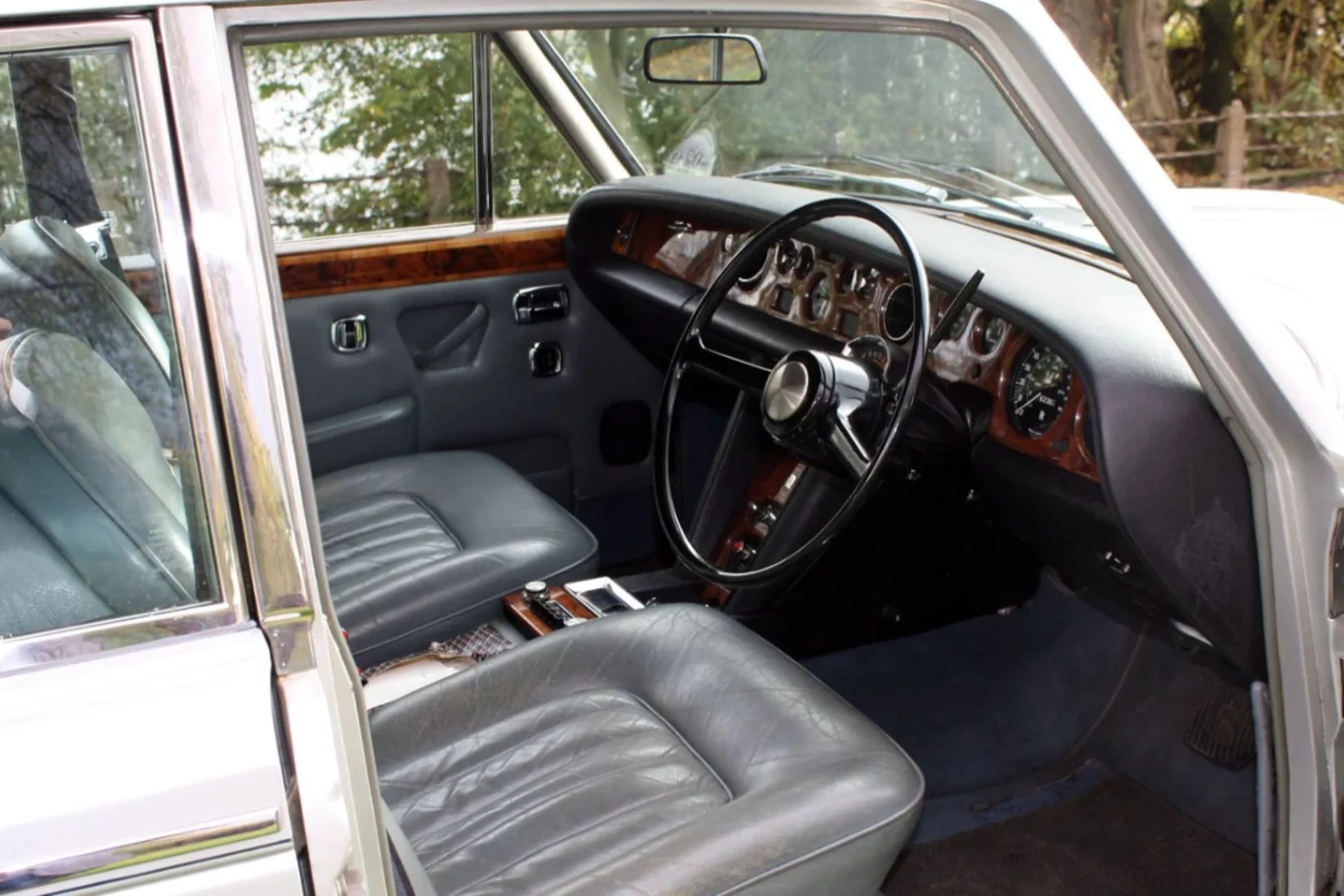 Tak wyglądało wnętrze Rolls Royce'a Freddiego Mercurego. Luksusowe wnętrze klasycznego samochodu z drewnianymi wykończeniami, starannie wyprofilowaną deską rozdzielczą, dużą kierownicą oraz wygodnymi, skórzanymi fotelami w kolorze szarym. Przez otwarte drzwi widać fragment przyrody za pojazdem.