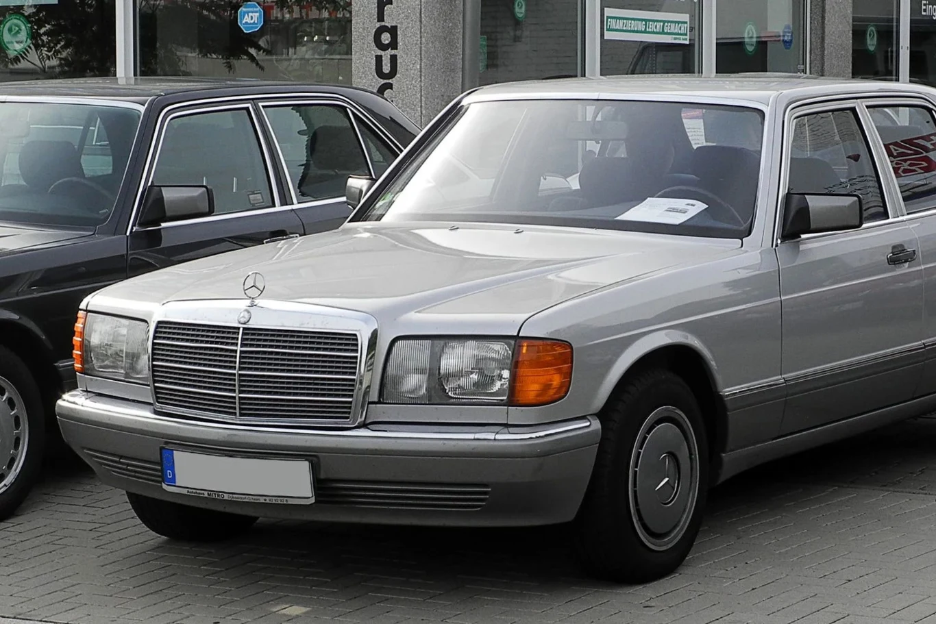 Mercedes 420 SEL towarzyszył Freddiemu Mercuremu do ostatnich dni. Srebrny sedan marki Mercedes-Benz ustawiony na parkingu, zaparkowany obok innego pojazdu, widoczne duże reflektory, klasyczna krata chłodnicy z logo na masce oraz dobrze zachowane nadwozie.