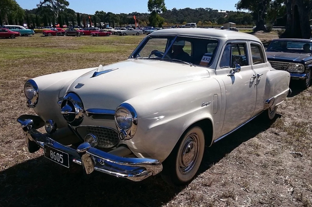 Studabaker Champion był wyjątkową perełką w kolekcji Freddiego. Biały klasyczny samochód osobowy Studebaker Commander Champion z lat 50-tych, zaparkowany na trawiastej powierzchni podczas zlotu zabytkowych pojazdów, w tle widoczne inne stare samochody oraz drzewa.