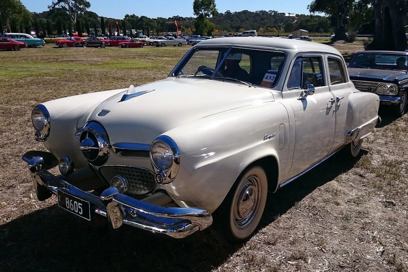 Studabaker Champion był wyjątkową perełką w kolekcji Freddiego. Biały klasyczny samochód osobowy Studebaker Commander Champion z lat 50-tych, zaparkowany na trawiastej powierzchni podczas zlotu zabytkowych pojazdów, w tle widoczne inne stare samochody oraz drzewa.