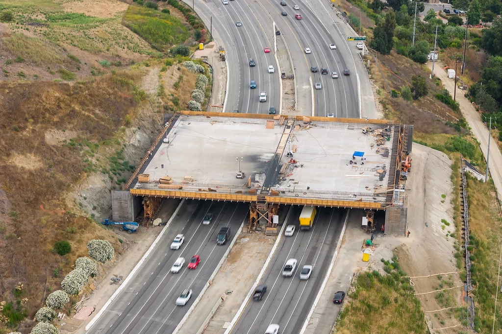 Docelowo obiekt będzie przebiegał nad dziesięciopasmową autostradą. Rozbudowa wiaduktu nad autostradą, gęsty ruch samochodów pod prowizoryczną konstrukcją, w tle zieleń oraz poboczne drogi techniczne.