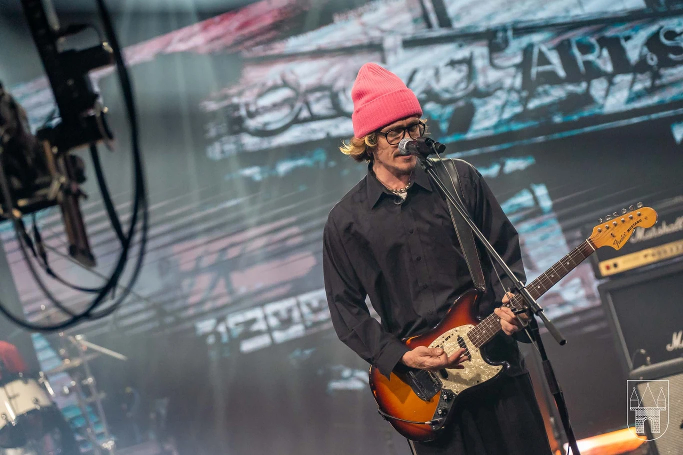 Zespół LÖS na scenie Wielkopolskich Rytmów Młodych Muzyk w różowej czapce śpiewający do mikrofonu i grający na gitarze elektrycznej na scenie koncertowej, w tle widać elementy oświetlenia i fragmenty perkusji.