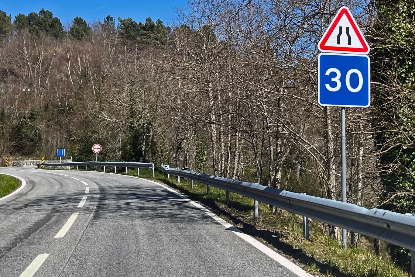 Ten znak zaskakuje kierowców. Co oznacza liczba na niebieskim tle? Zakole drogi prowadzącej przez zalesioną okolicę, przy której ustawiono znaki drogowe informujące o zwężeniu jezdni oraz ograniczeniu prędkości do 30 km/h, metalowe bariery ochronne wzdłuż pobocza, bez widocznych pojazdów i przechodniów.