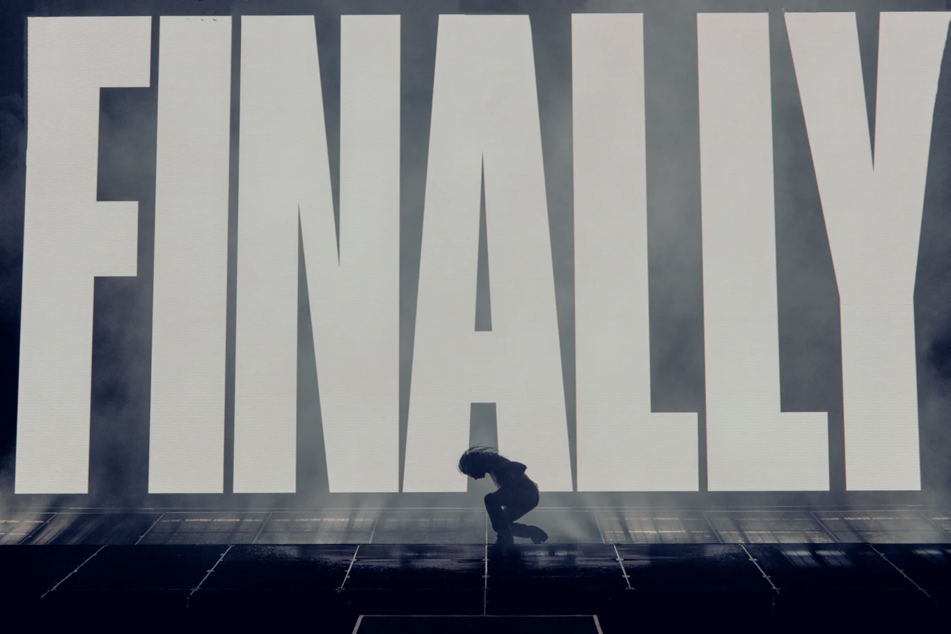 Duży napis FINALLY dominuje tło sceny, na której na pierwszym planie osoba przyklęka w lekkim cieniu i półmroku.