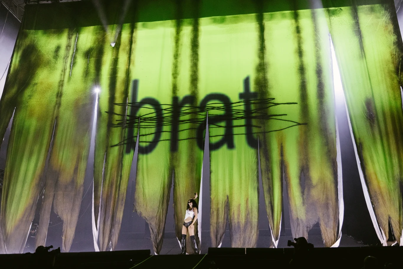 Osoba stojąca na scenie z dużymi zielonymi kotarami, na których widnieje czarne, przekreślone słowo 'brat', tło podświetlone jest efektami świetlnymi.