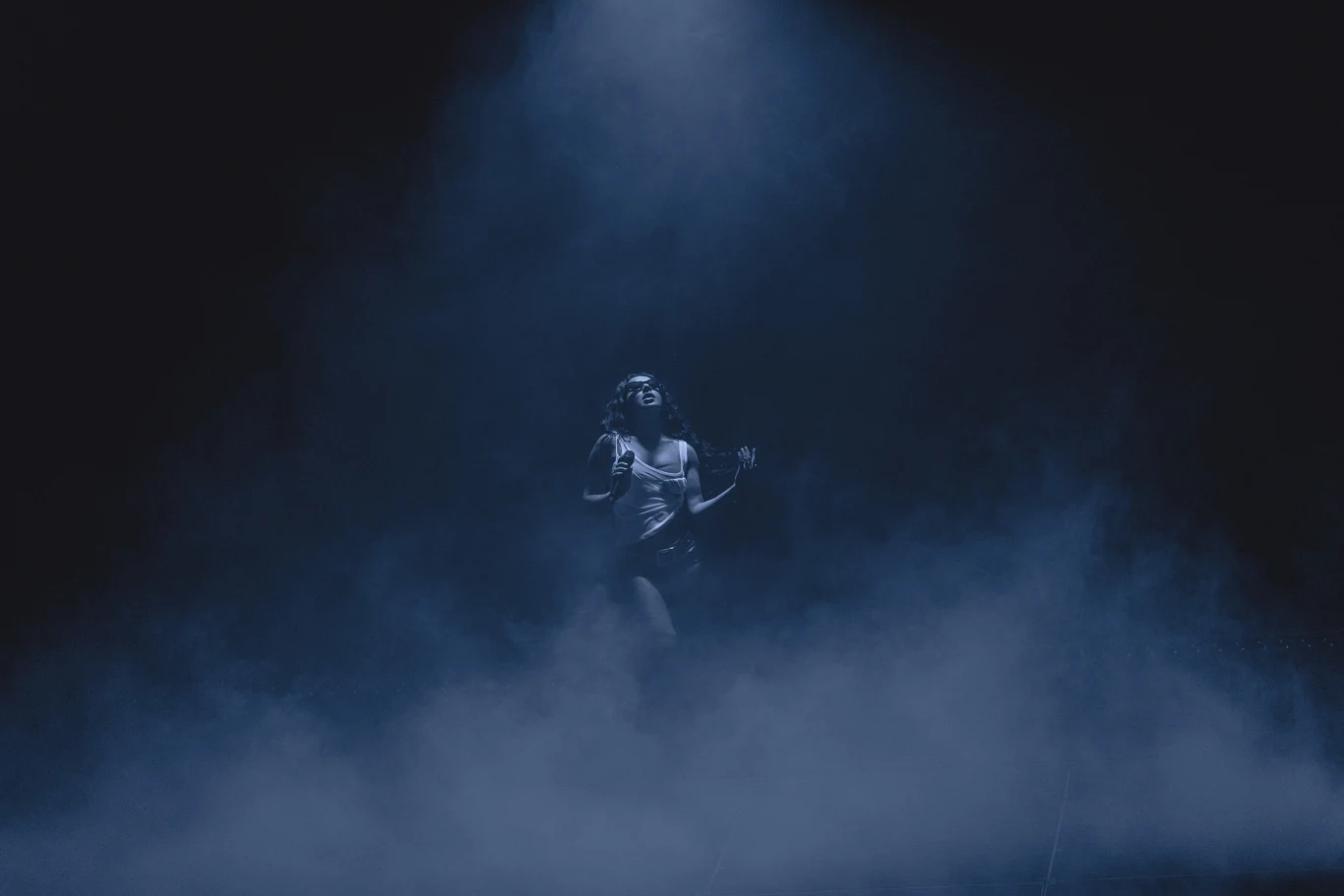 Kobieta w sportowym stroju stoi na scenie otulona mgłą i oświetlona jasnym światłem padającym z góry, wokół panuje ciemność tworząca tajemniczy nastrój.