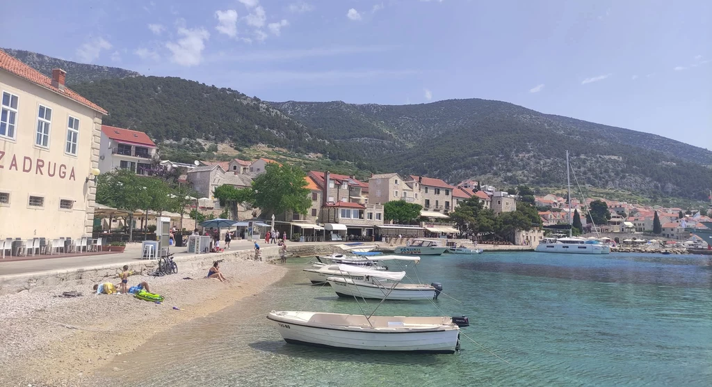 Bol w Chorwacji Plaża nad brzegiem morza z kilkoma łodziami zacumowanymi przy brzegu, w tle zabudowania małego miasteczka oraz góry pod niebieskim, lekko zachmurzonym niebem.