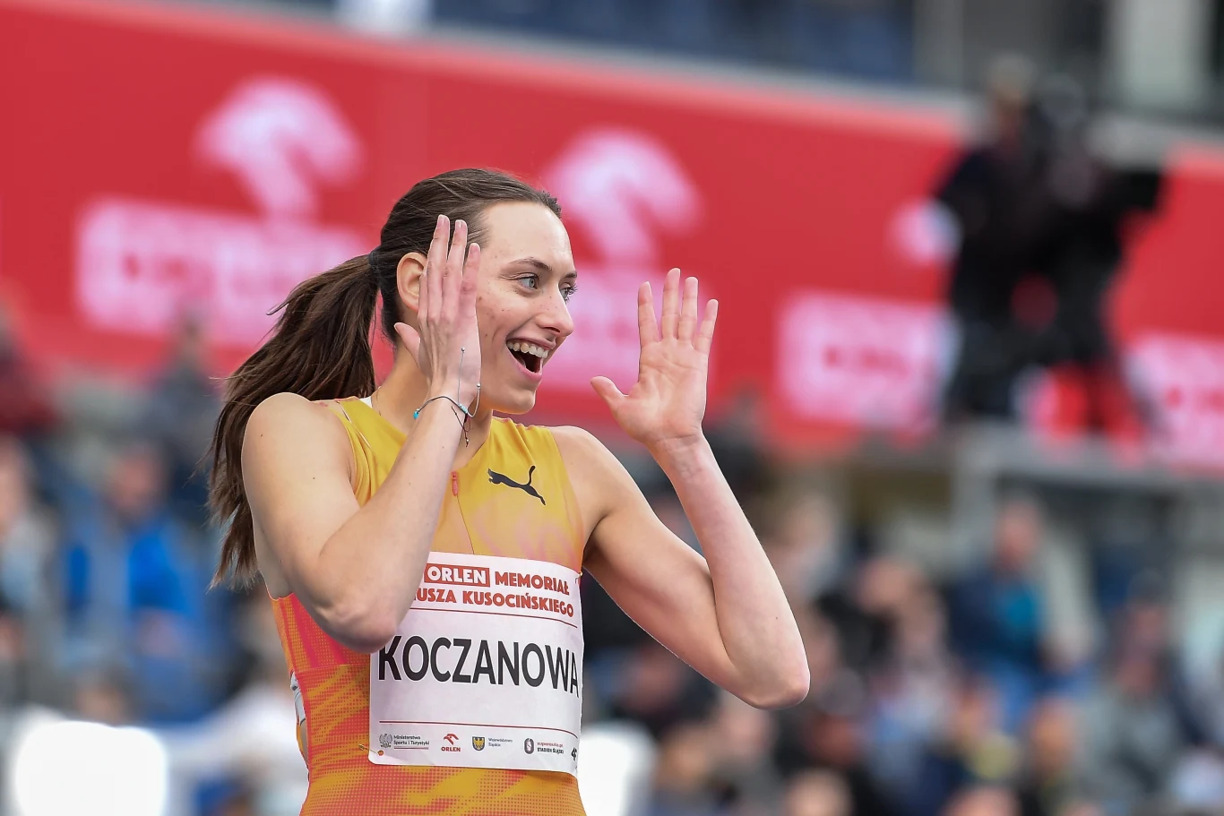 Zawodniczka w sportowej koszulce z numerem startowym podczas wydarzenia lekkoatletycznego, uśmiechnięta, wykonująca gesty dłońmi, w tle rozmyte sylwetki widzów i czerwone banery reklamowe.